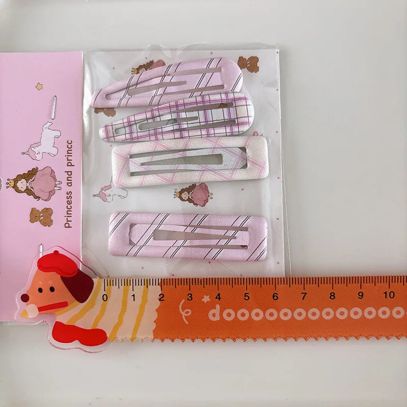 Four pink hair clips with plaid patterns in a clear plastic sleeve, shown next to a pink branded card and an orange ruler on a white background.
