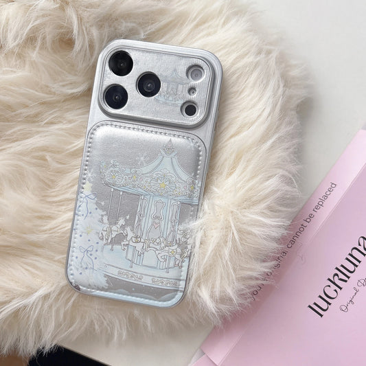 A silver phone case with a decorative carousel illustration, displayed on a fluffy cream faux-fur surface next to a pink book.