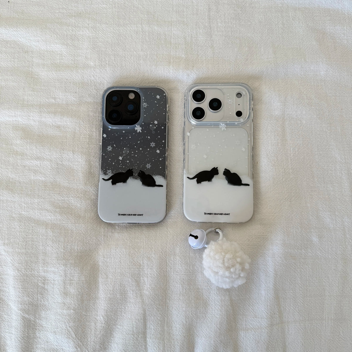Two clear phone cases with a winter snow design and black cat silhouettes lying on a textured off-white fabric surface. One case is dark gray with white snowflakes, and the other is clear with a white lower section.