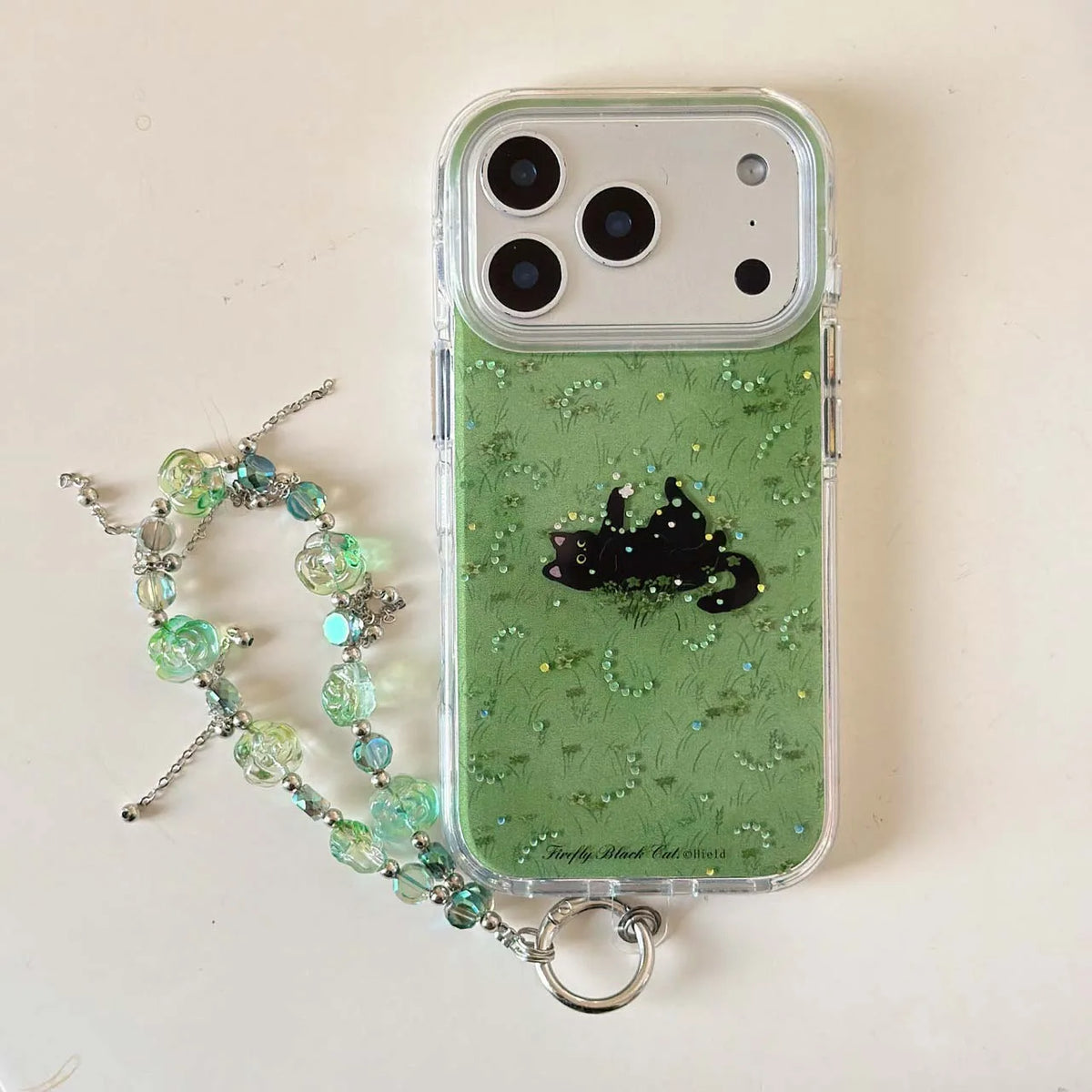 A clear phone case with a green glittery interior, a black cat silhouette design, and a matching beaded wrist strap with a silver ring, displayed on a light beige surface.