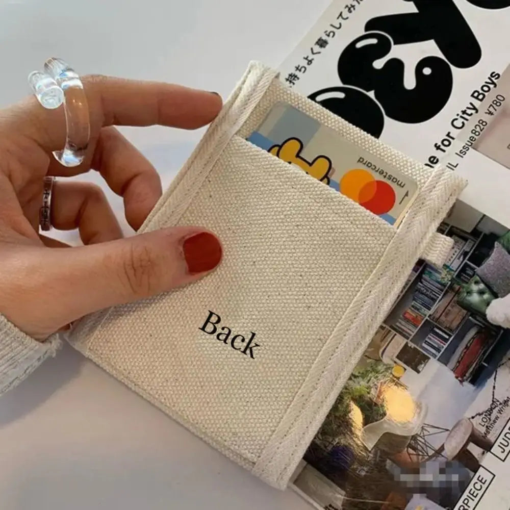 A close-up of a hand holding a small canvas card holder or wallet in cream/off-white with the word 'Back' printed on it. The holder contains cards, and part of a magazine or book with bold black text is visible behind it.