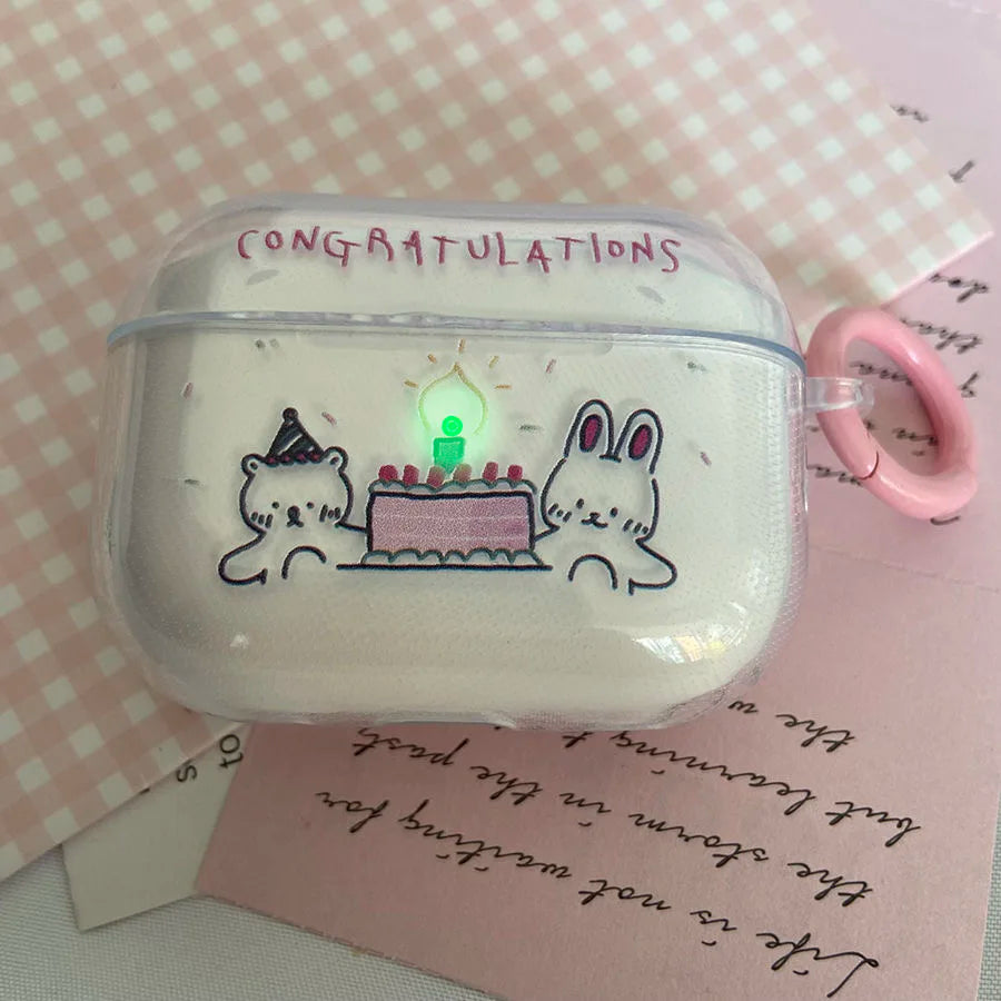 A clear plastic AirPods case cover with a cute birthday illustration of a bear and a bunny around a cake, featuring a pink 'CONGRATULATIONS' text and a pink carrying loop, resting on pink patterned paper.