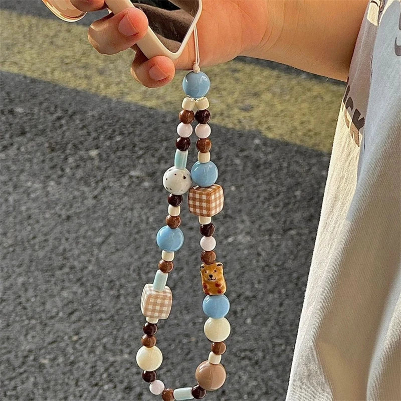 A close-up of a hand holding a phone case with a long decorative beaded strap outdoors. The strap features pastel blue, brown, cream, and checkered beads, along with small bear and dot charms.