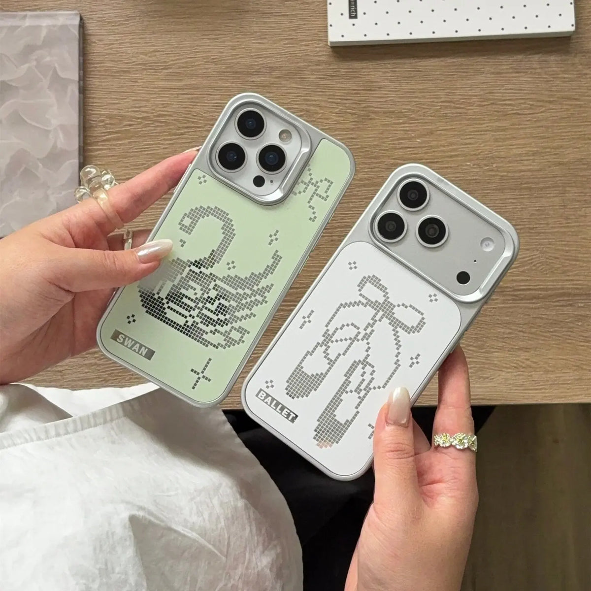 Two phone cases held in hands over a wooden desk. One case is pale mint green with a pixelated swan design, and the other is light gray with a pixelated bow design.