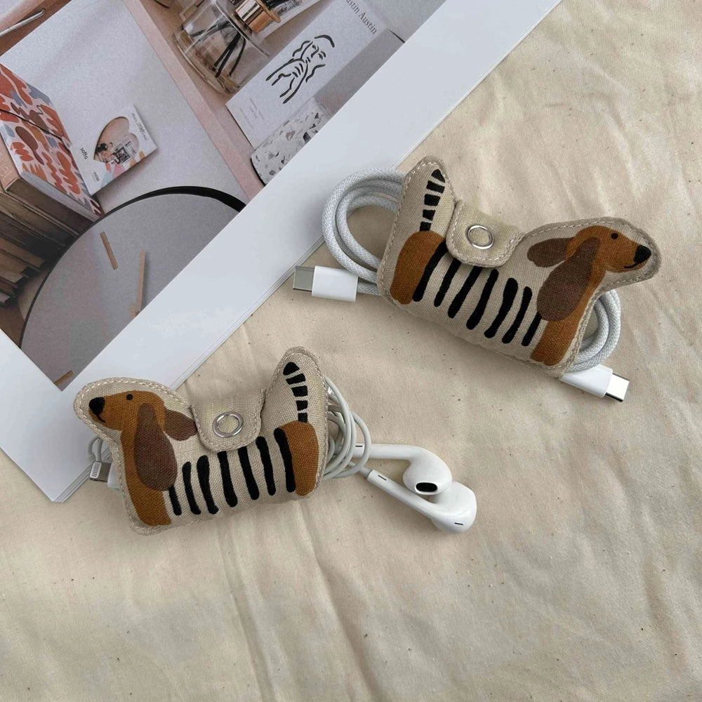 Two dachshund-shaped cable organizers made of beige faux leather with black stripes and brown ears, shown holding white earphones and charging cables on a beige fabric surface with a magazine in the background.