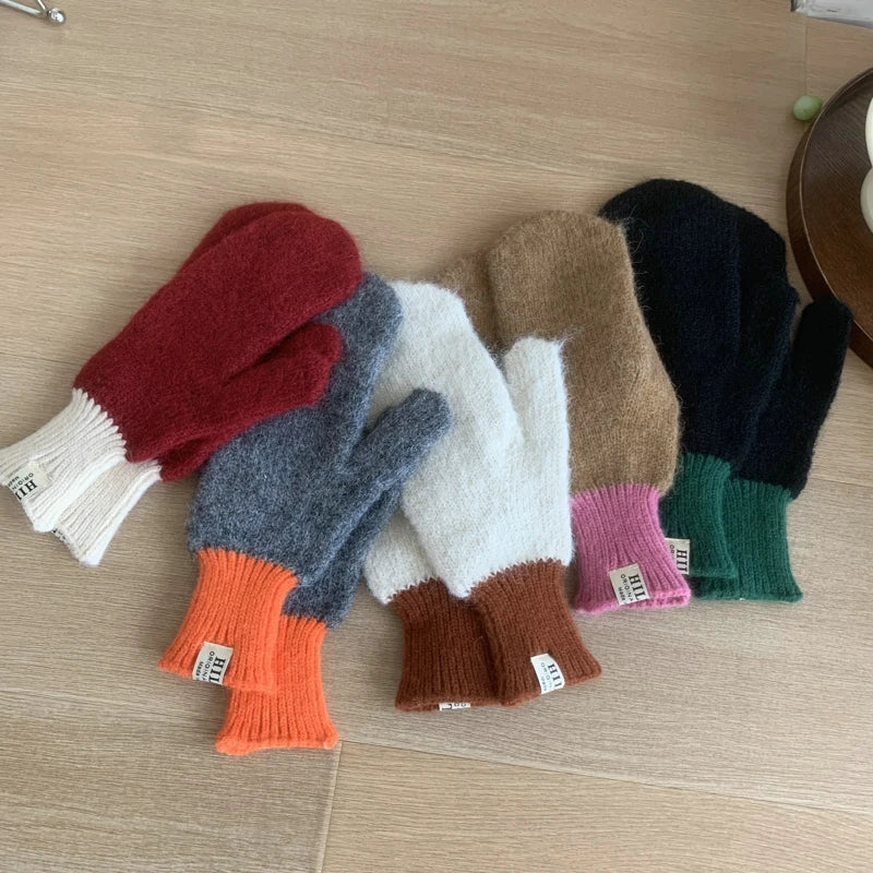 Five pairs of fuzzy knit winter mittens in assorted colors are arranged on a light wood floor. The colors include red with white, gray with orange, white with brown, tan with pink, black with green, and a dark navy with green cuff.