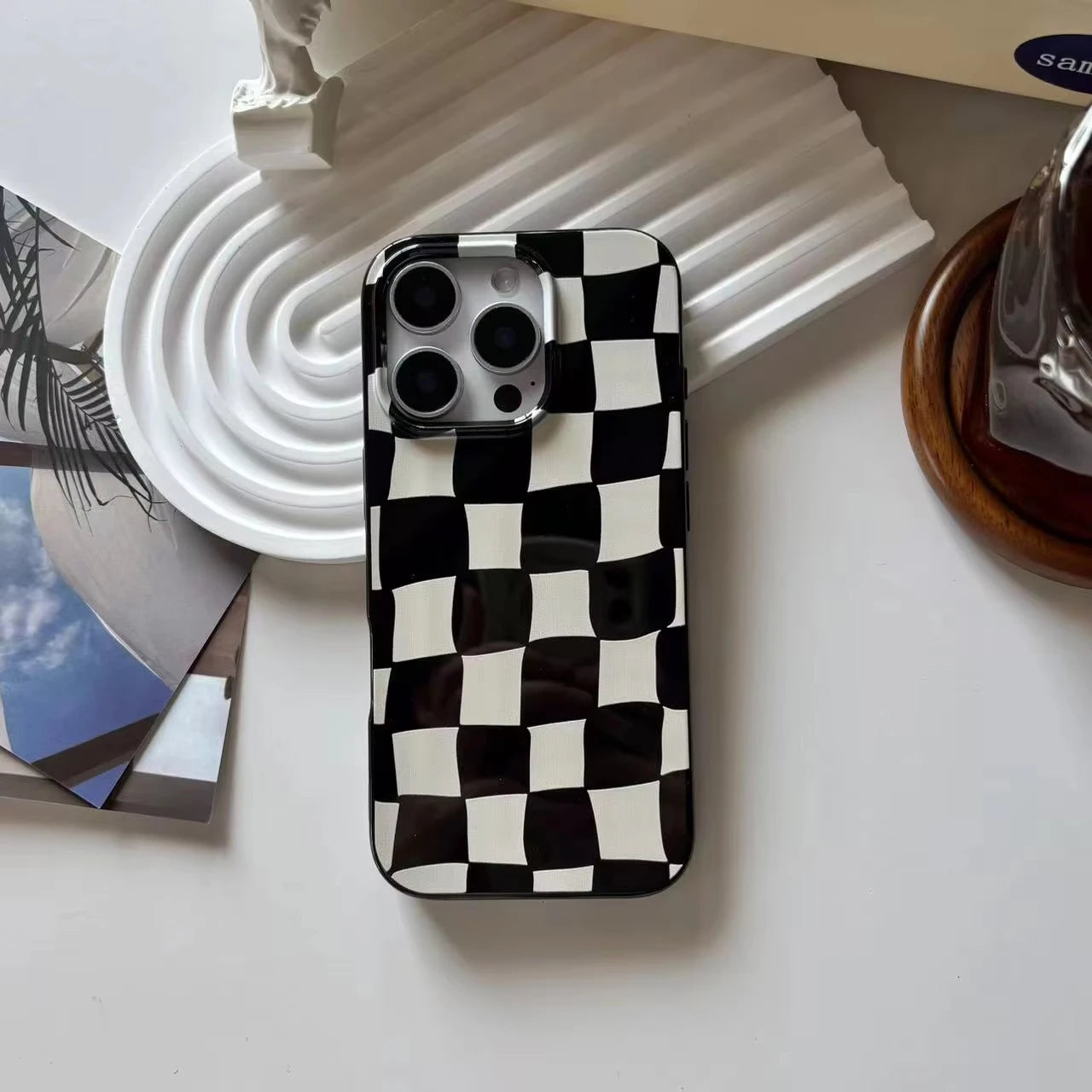 A phone case with a black and white checkerboard pattern, shown on a white surface with decorative props including a white pleated plate and a glass object.