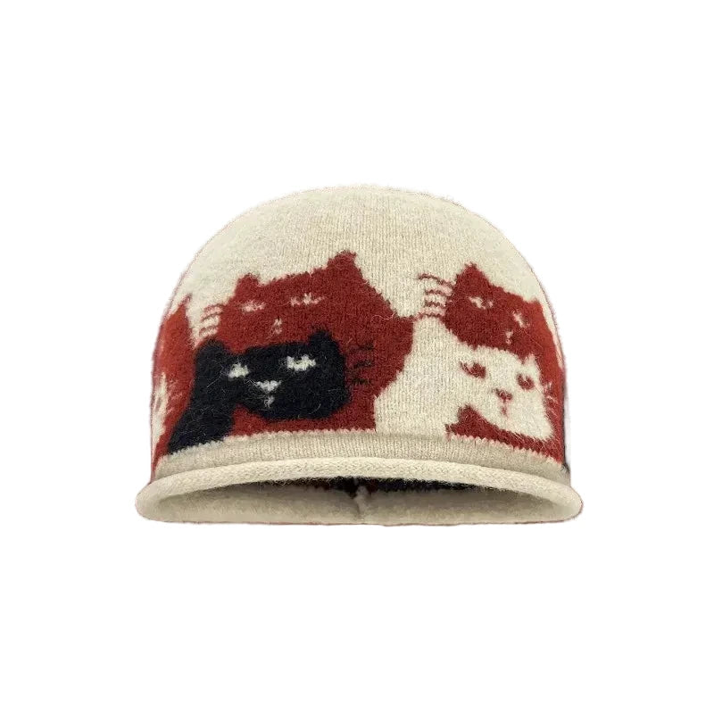 A knit beanie hat from Coisini Studio featuring a pattern of stylized cat faces in red, cream, and black. The hat is centered against a solid dark red background with white text at the top and bottom.