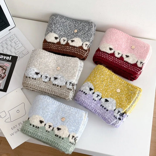Five folded chunky knit scarves with sheep patterns in different colors (gray, brown, light gray, pink, red, yellow, lavender, and mint green) arranged on a white desk next to a laptop and a notebook.