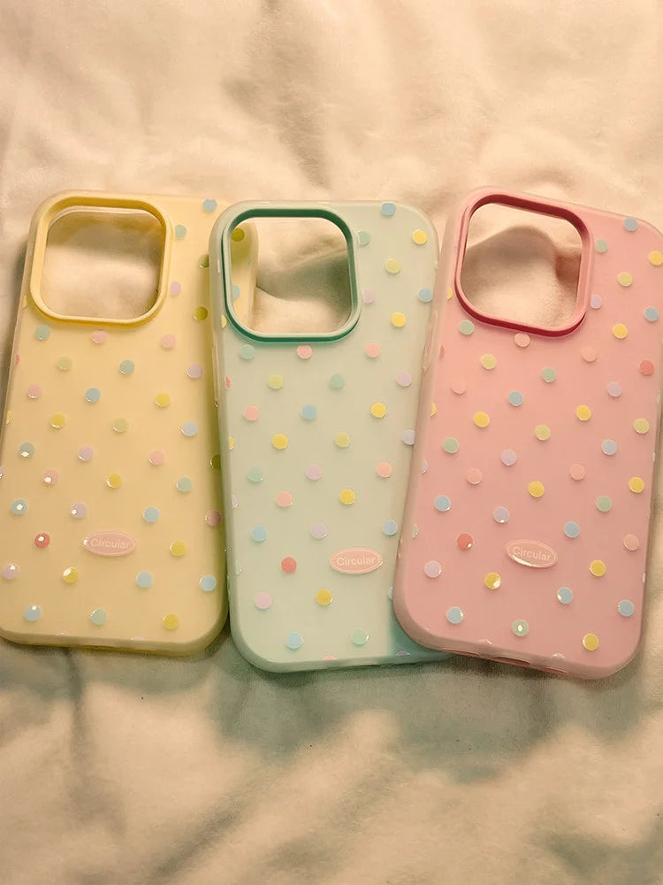 Three pastel-colored phone cases with a polka dot pattern, displayed side-by-side on a soft, light-colored fabric background.