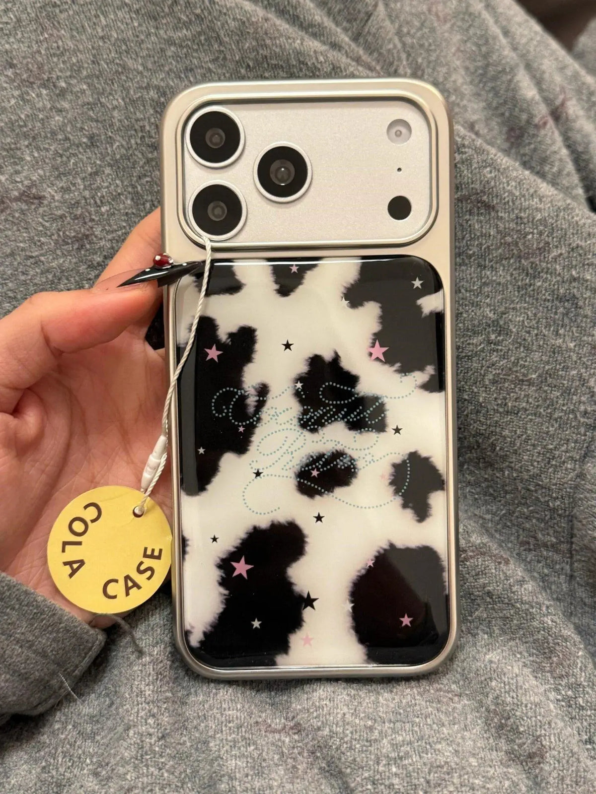 A phone case with a black and white cow print pattern, decorated with small pink stars and a silver glitter script logo, held by a hand with a yellow 'COLA CASE' tag against a gray knit background.