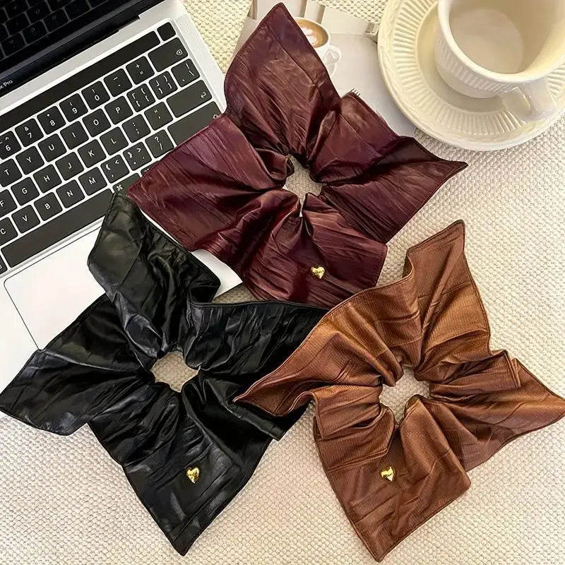 Three large satin-like scrunchies in black, deep burgundy, and warm brown, each with a small gold heart detail, arranged on a textured beige surface next to a laptop and a cup on a saucer.