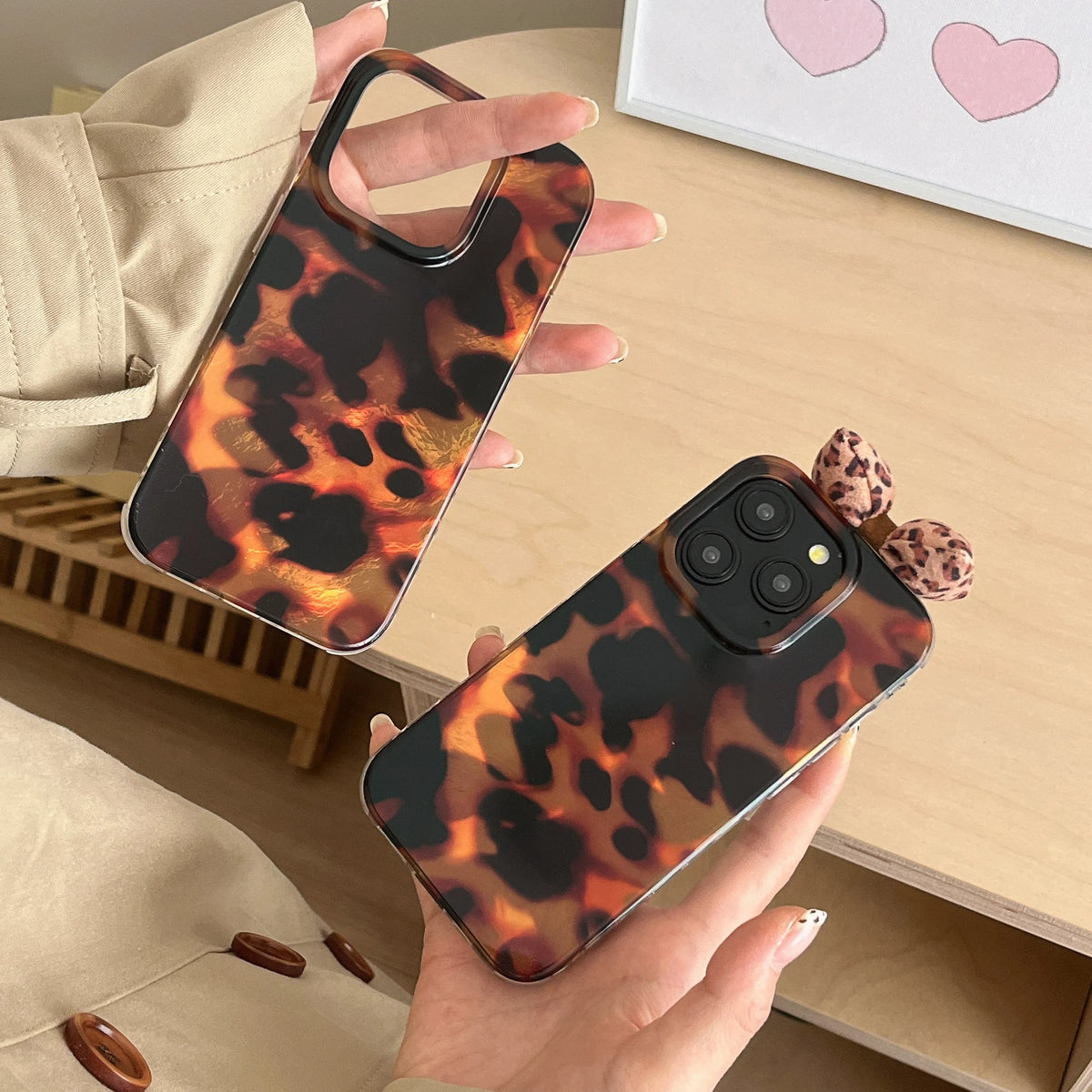 Two leopard print phone cases are shown, one held by a hand and one resting on a wooden table. The phone case features a warm brown, black, and orange leopard pattern, with one case styled with a small matching leopard bow on the side.