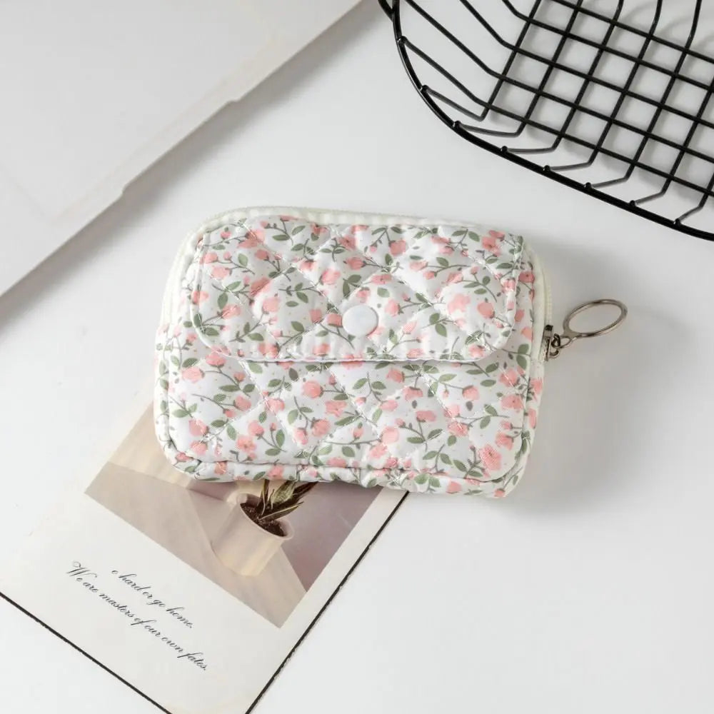 A quilted coin purse with a pink and green floral pattern, featuring a snap button closure and a metal key ring, styled on a white surface with a magazine and a black wire basket.