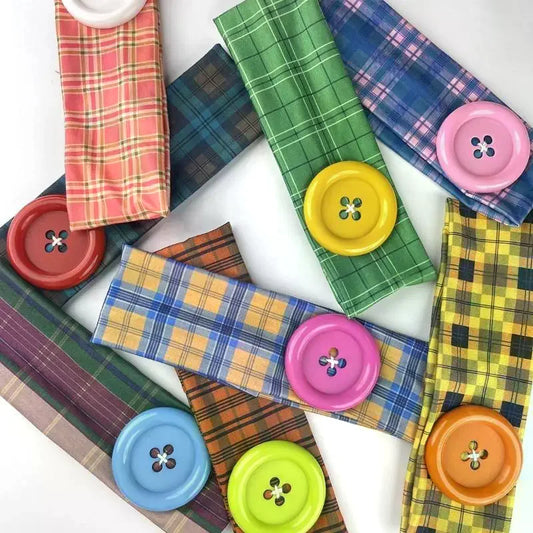 A flat-lay arrangement of colorful plaid and checkered fabric headbands, each placed on top of a large decorative button in different colors including pink, green, blue, yellow, orange, and red, on a white background.