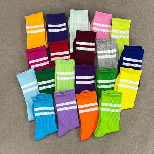 A collection of colorful athletic socks arranged in a pyramid on a textured gray fabric background. Each sock features two white horizontal stripes near the top, with various colors including yellow, blue, pink, green, orange, purple, and red.