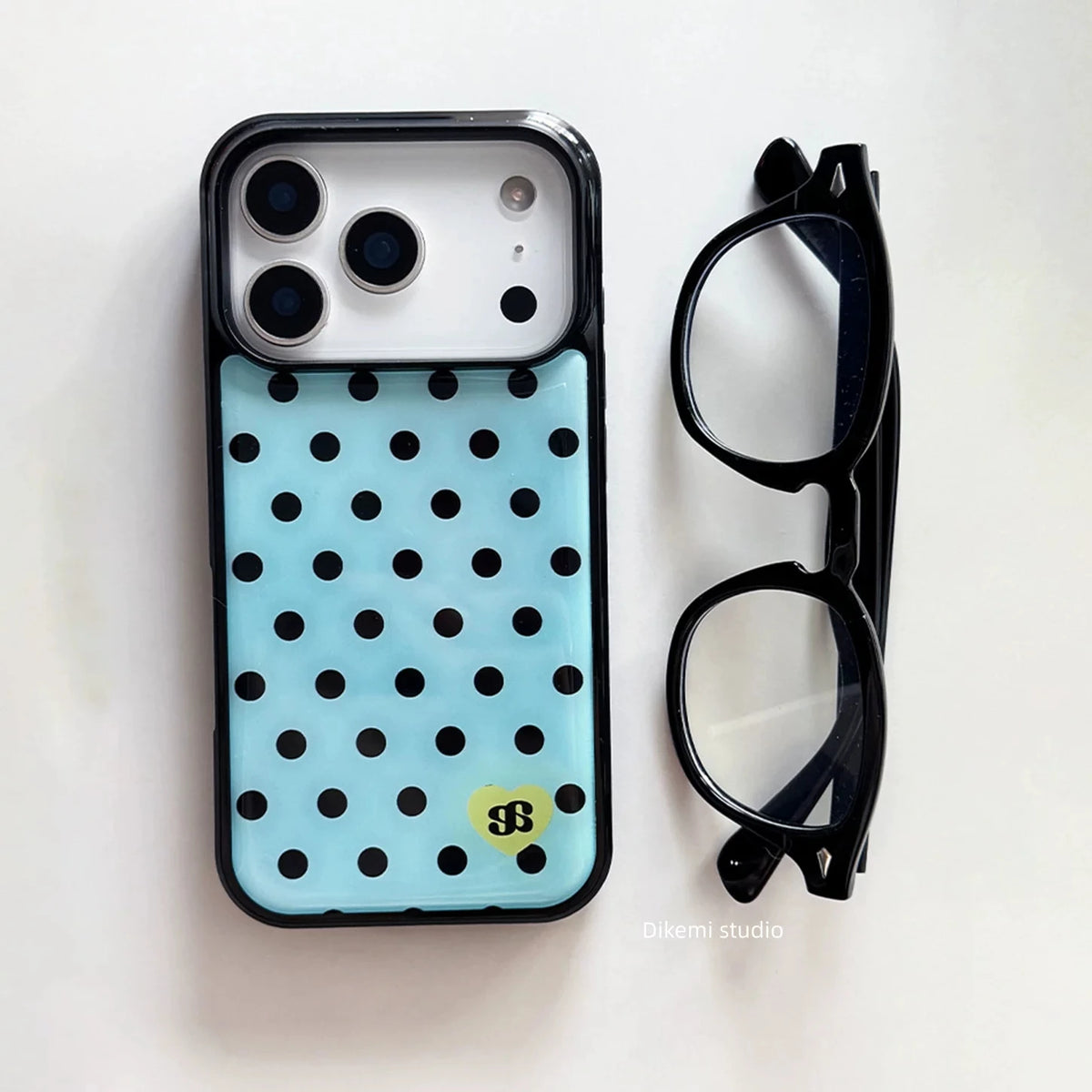 A light blue phone case with black polka dots and a small yellow dollar-sign logo, shown next to a pair of black eyeglasses on a white background.