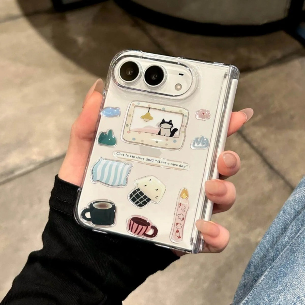 A hand holding a clear phone case decorated with cute pastel stickers, including a cat in a bed, pillows, a cup of coffee, and a candle, shown against an indoor background.