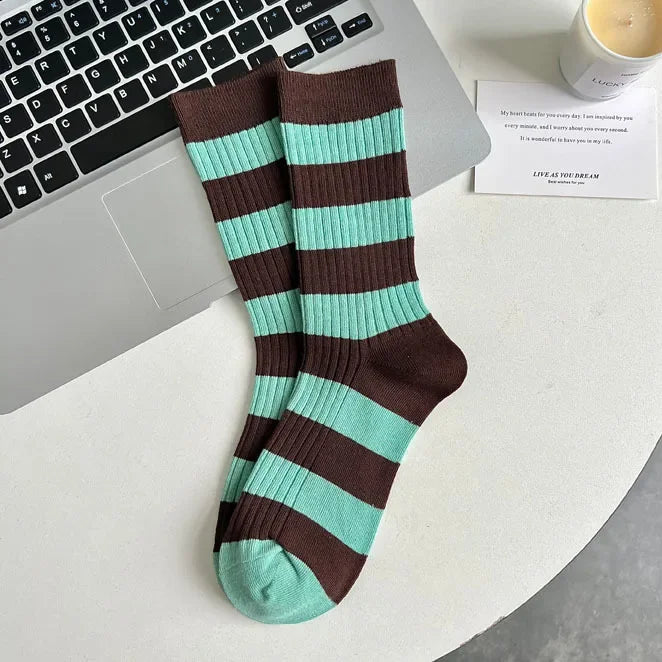 A pair of ribbed crew socks with alternating dark brown and mint green horizontal stripes, styled on a white desk next to a laptop, a candle, and a small card.