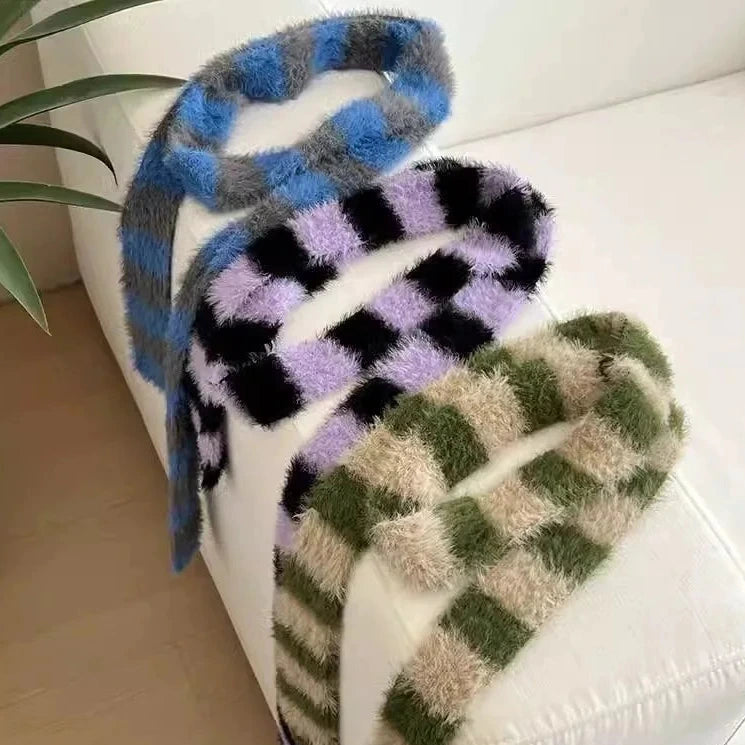 Three fuzzy striped knit scarves draped over a white sofa: blue and gray, black and lavender, and olive green and beige. A plant leaf is visible at the top left.