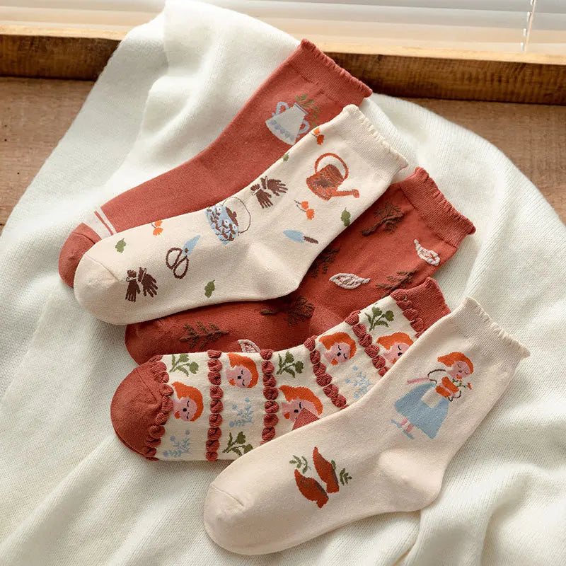 Three pairs of patterned ankle socks arranged on a soft white fabric surface. The socks feature whimsical autumn and folk-art illustrations including a teapot, mushrooms, a watering can, leaves, and a girl in a blue dress, with warm rust and cream tones throughout.