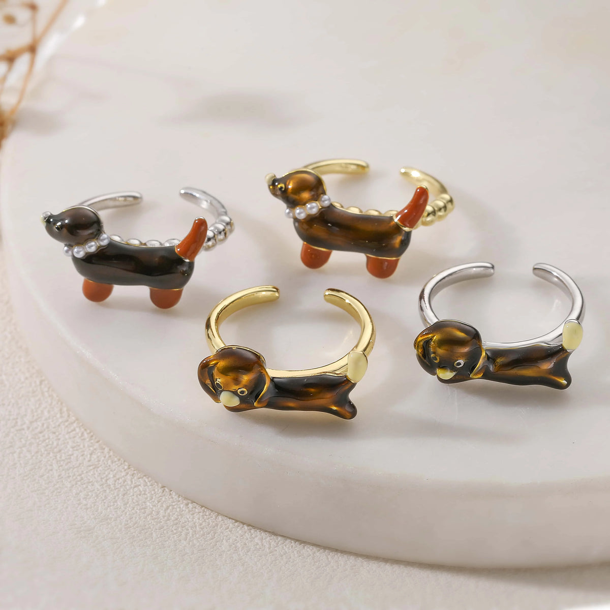 Four dog-shaped ring earrings displayed on a white marble tray. The earrings feature glossy brown and black enamel bodies, orange tails and legs, and are finished in three metal colors: gold, silver, and rose gold.