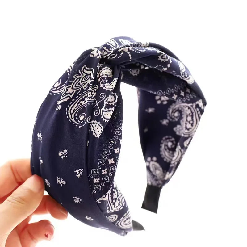 A navy blue and white paisley bandana headband being held by a hand against a plain white background.