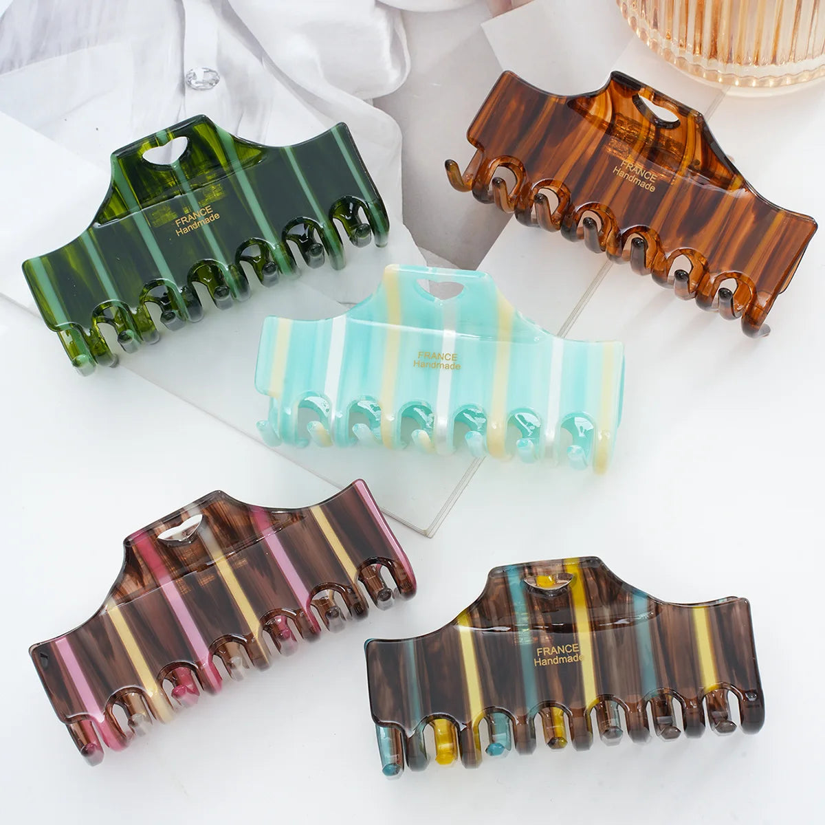 Five hair claw clips in assorted marbled and striped acrylic designs, including green, brown, light blue, pink, and multicolor patterns, arranged on a white surface with a glass plate and white fabric in the background.