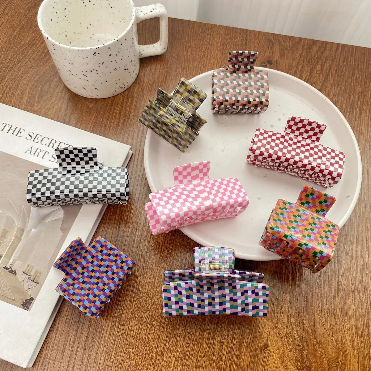 Six claw hair clips with different colorful geometric patterns are arranged on a white plate and a wooden table next to a speckled mug and a magazine.