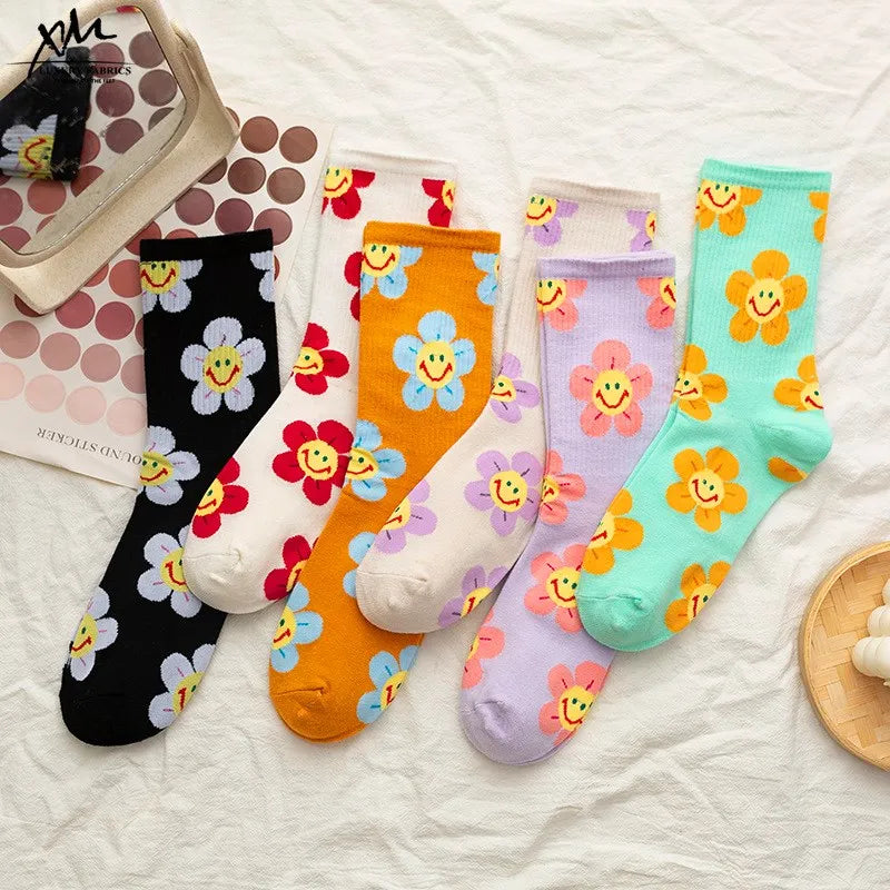 Five pairs of crew socks with colorful smiling flower patterns are arranged on a white fabric surface. The socks come in black, white, orange, lavender, and mint green, each decorated with different flower colors and yellow smiley faces.