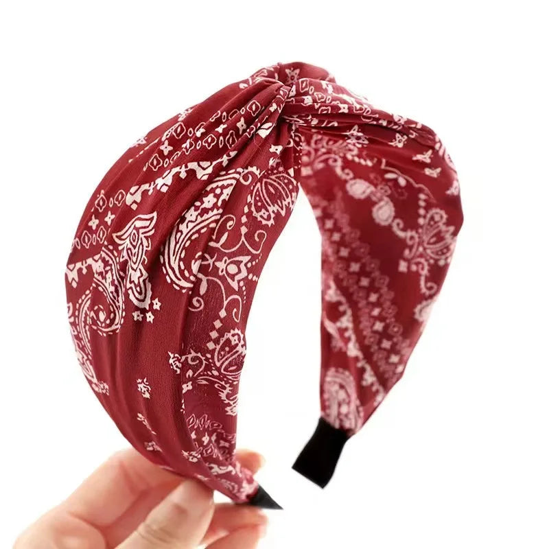 A red fabric headband with a white paisley bandana pattern, held by a hand against a plain white background.