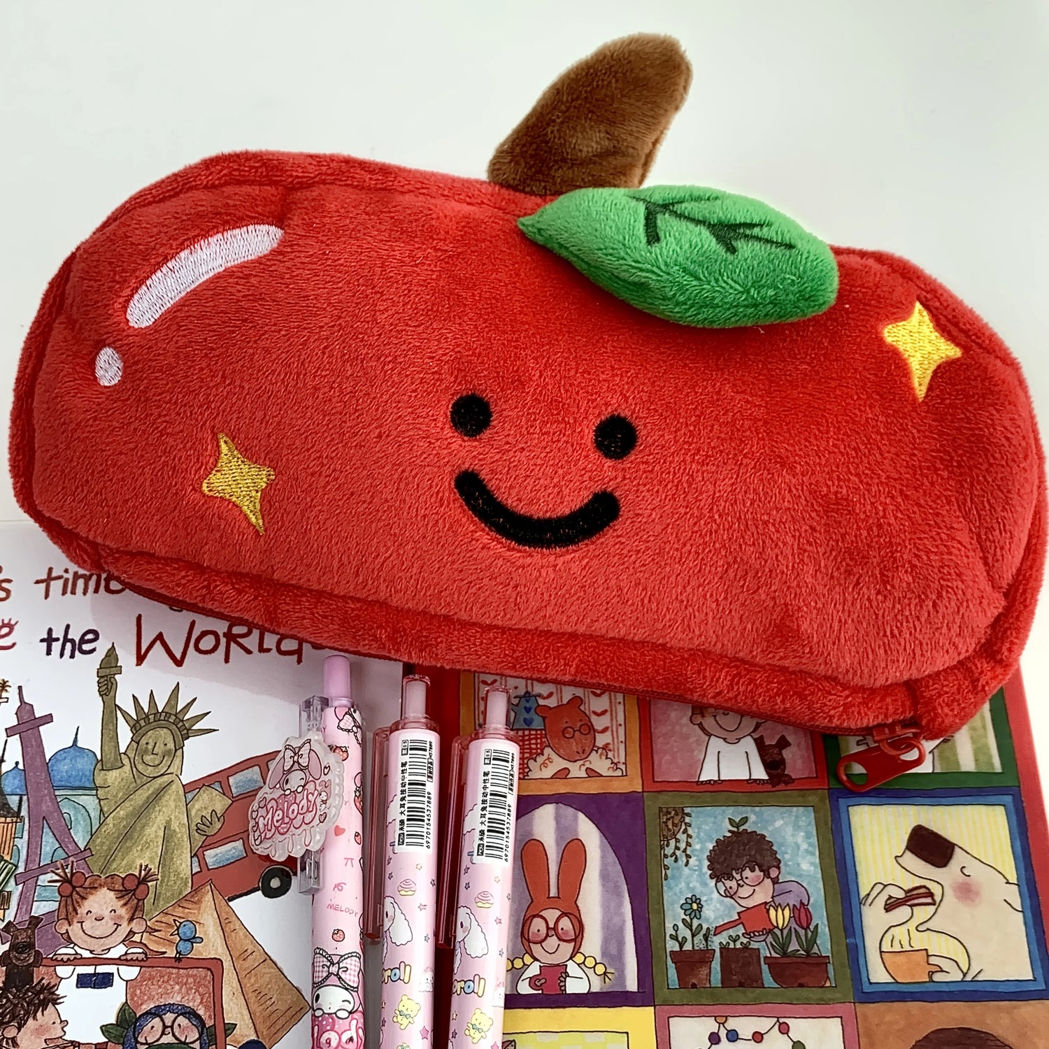 A red plush apple-shaped pencil case with a smiling face, green leaf, and yellow star details, shown with three pink patterned pens on top of a colorful illustrated card.