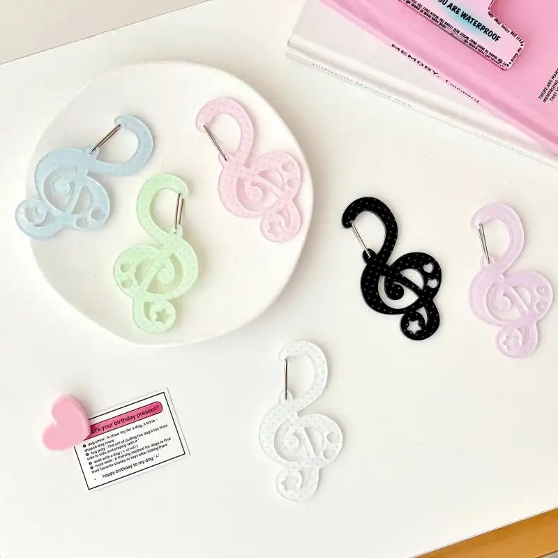 A styled flat-lay of six treble clef keychain earrings in pastel and solid colors, arranged on a white plate and scattered on a white surface next to a pink book and a small pink card.