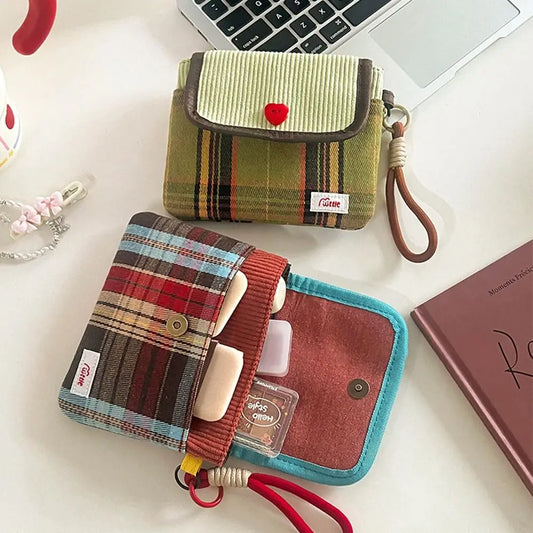 Two small fabric card holder wallets with plaid patterns are arranged on a white desk. The top wallet has a striped flap with a small red heart and brown trim, and the bottom wallet has a red-and-blue plaid body with a red elastic strap and card slots.