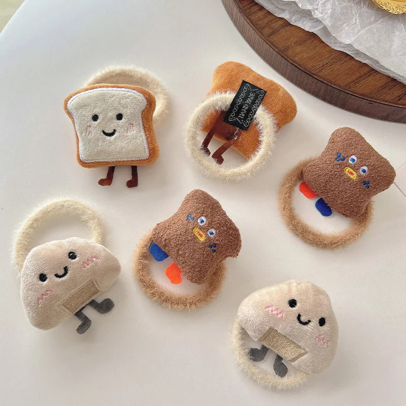 Five cute plush hair ties shaped like toast, muffins, and a mushroom, arranged on a white surface.