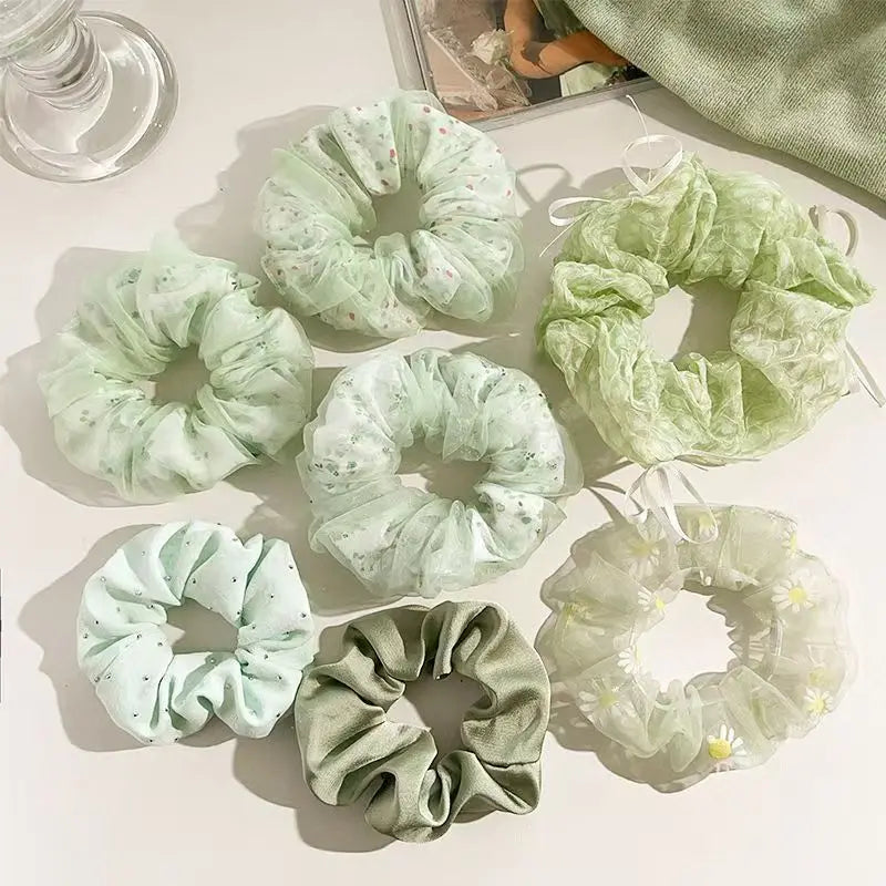 Seven sheer scrunchies in soft pastel shades are arranged on a white surface. The set includes different fabrics and prints: floral mesh, dotted tulle, and satin-like textures in mint green, pale pink, light blue, sage green, and white with small yellow daisies.