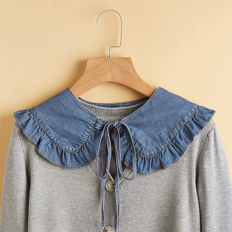 A gray knit cardigan with a large blue denim ruffled Peter Pan collar and tie detail, hanging on a wooden hanger against a warm beige background.