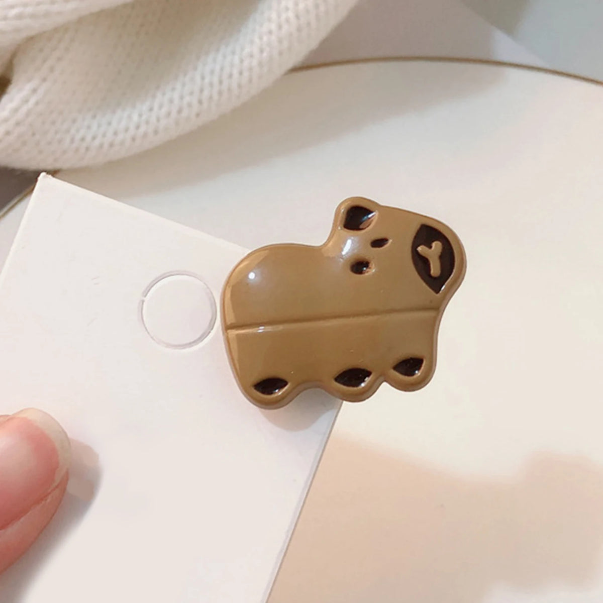 A small glossy brown capybara-shaped accessory resting on a white card, with a finger holding the card and a soft white fabric in the background.