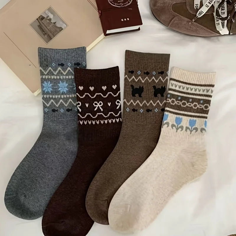 Four pairs of patterned crew socks laid flat on a white surface: one heather gray with blue and white Nordic motifs, one dark brown with white heart and bow patterns, one taupe-brown with black dog silhouettes, and one cream with blue and brown floral/Fair Isle patterns.