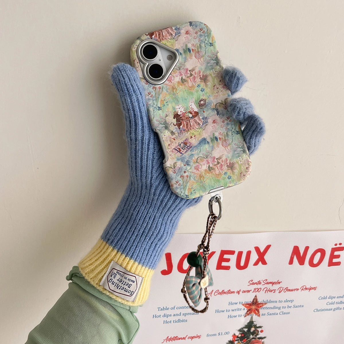A hand wearing a blue and yellow knit glove holds a phone with a pastel floral phone case and a dangling ornament keychain against a plain off-white wall.