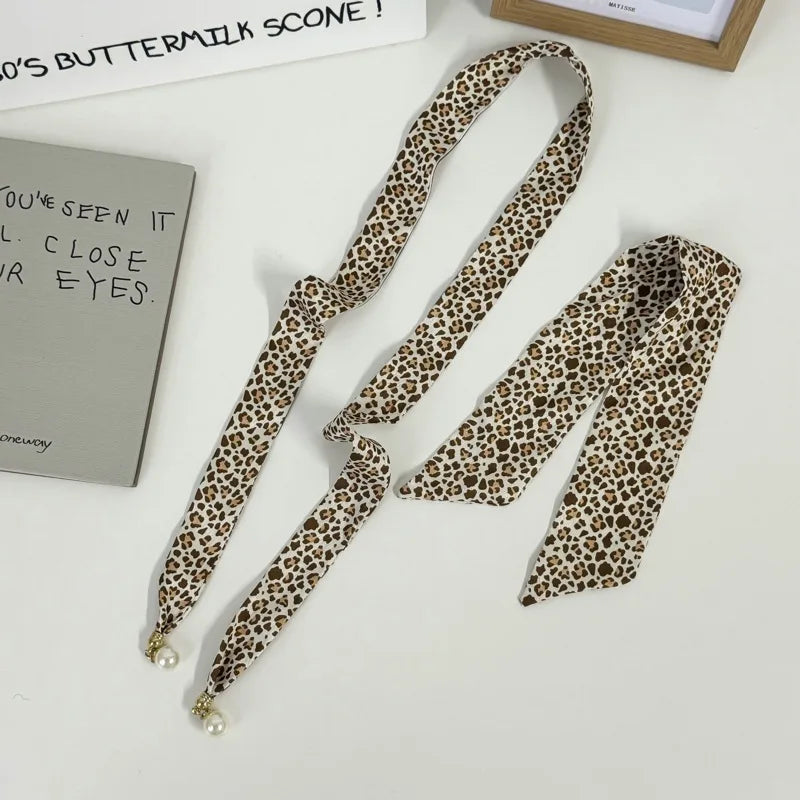 A leopard print necktie with pearl-like tips, displayed on a white surface alongside decorative props including a notebook, a book, and a small wooden frame.