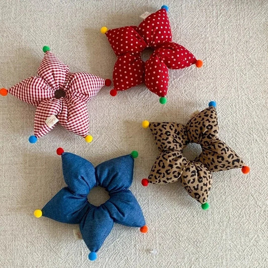 Four fabric flower-shaped scrunchies arranged on a textured off-white background. The set includes a red and white gingham flower, a red and white polka dot flower, a blue solid-color flower, and a leopard print flower, each with colorful pom-pom ends.