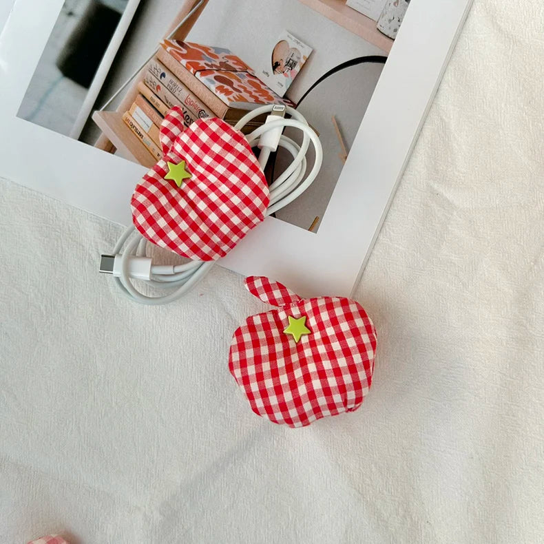 Two red and white gingham apple-shaped cable organizers with small yellow star accents, shown holding white charging cables on a white fabric surface next to a picture frame.
