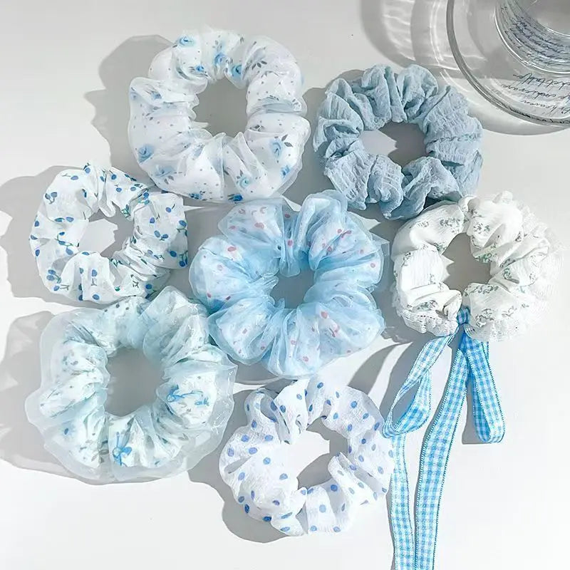 Six light blue and white scrunchies with various patterns (dots, lace, gingham ribbon) arranged on a white surface.