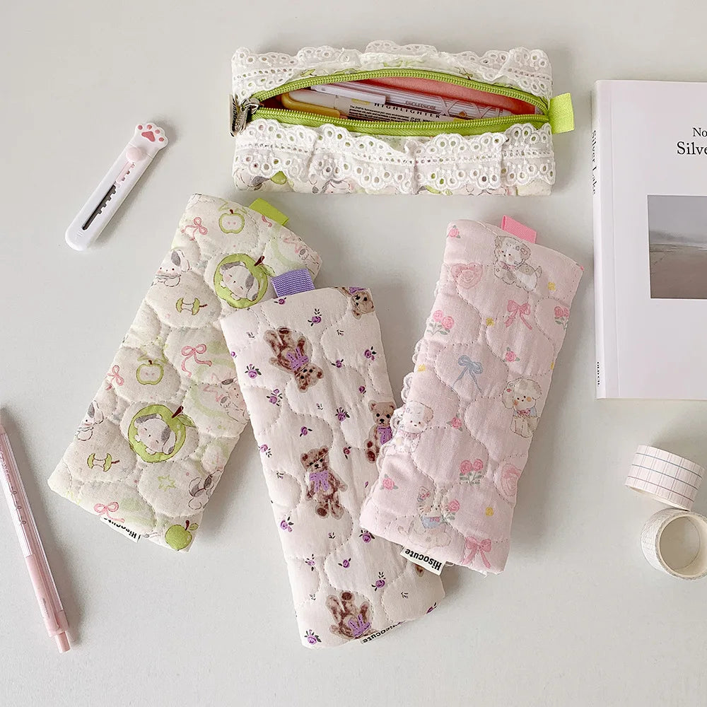 Three quilted pencil cases with cute animal prints are arranged on a light gray surface alongside stationery props, including a white lace zip pouch, a pink pen, a utility knife, and a book.