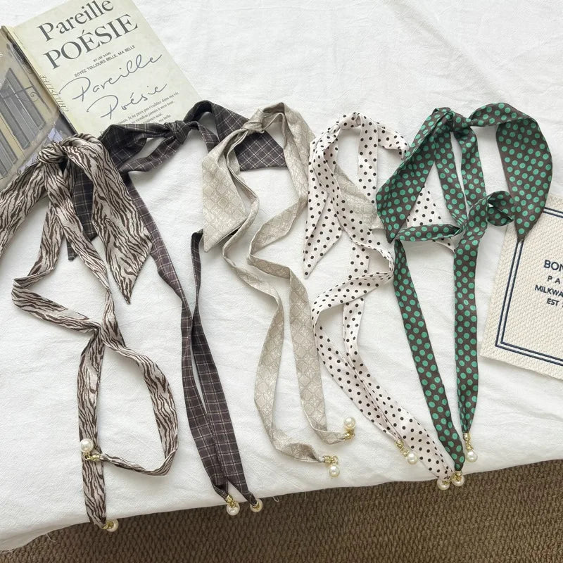 Four long satin ribbon ties with pearl beads at the ends are arranged on a white fabric surface. The ribbons have different patterns: a gray-and-white abstract print, a dark gray plaid, a light beige micro-dot pattern, and a green-and-white polka dot.
