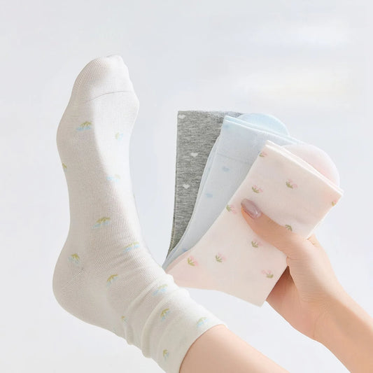 A person wearing a white ankle sock with small pastel rainbow prints, while holding a stack of folded socks in soft pastel colors against a clean white background.
