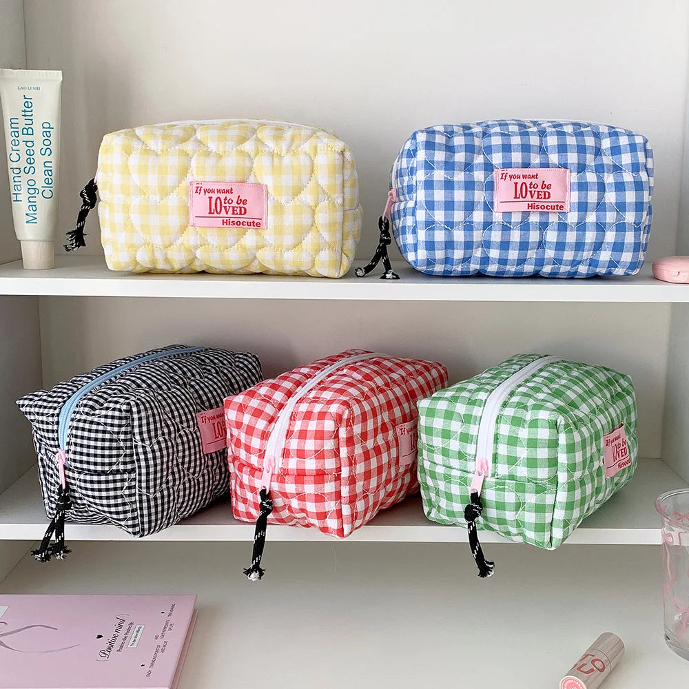 Four quilted cosmetic pouches with gingham checks are displayed on white shelves. The top row has a yellow-and-white checkered pouch and a blue-and-white checkered pouch, both with pink 'If you love 10,000 loves' labels.