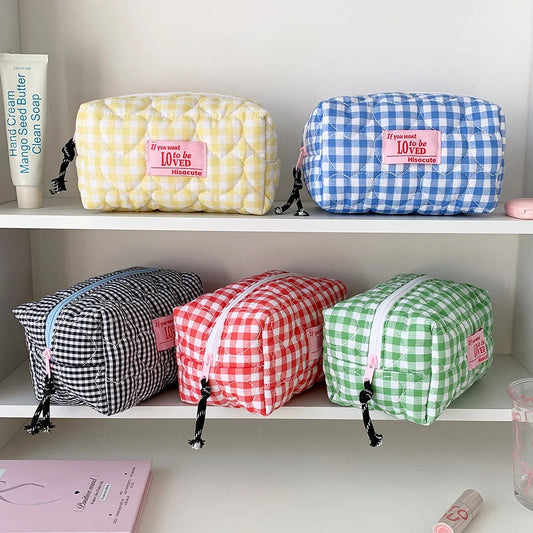 Four quilted cosmetic pouches with gingham checks are displayed on white shelves. The top row has a yellow-and-white checkered pouch and a blue-and-white checkered pouch, both with pink 'If you love 10,000 loves' labels.