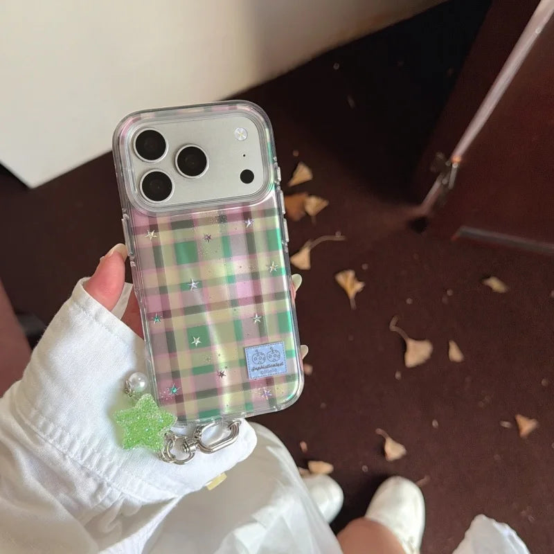 A hand holding a clear phone case with a pastel pink, green, and beige plaid pattern, decorated with small glitter stars and a dangling green glitter charm.
